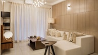 A modern, elegantly designed living room featuring a large white sectional sofa adorned with numerous decorative cushions. The room has a minimalist aesthetic with light wood-paneled walls and a sleek tile floor. A dark wooden coffee table, holding decorative objects, sits in front of the sofa. A contemporary tripod floor lamp adds warm lighting, complemented by overhead pendant lights. Large sheer curtains cover a window, creating a soft, diffused light throughout the space.