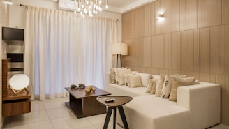 A modern, elegantly designed living room featuring a large white sectional sofa adorned with numerous decorative cushions. The room has a minimalist aesthetic with light wood-paneled walls and a sleek tile floor. A dark wooden coffee table, holding decorative objects, sits in front of the sofa. A contemporary tripod floor lamp adds warm lighting, complemented by overhead pendant lights. Large sheer curtains cover a window, creating a soft, diffused light throughout the space.