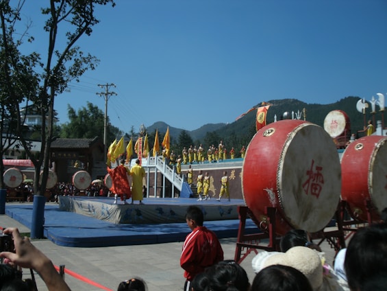 A lively outdoor performance featuring participants in yellow and red costumes on a raised platform with traditional Chinese drums in the foreground. Flags and banners add vibrant color to the scene, set against a backdrop of trees and hills under a clear blue sky. A crowd is gathered around, observing the event.