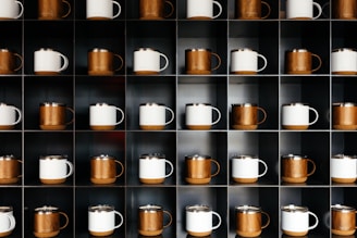 A set of four mugs arranged on a sleek black shelf, each showcasing different bold designs in black, white, and gold accents