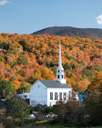 a white building with a steeple surrounded by trees and a hill with orange and yellow leaves