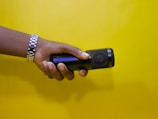 A hand wearing a silver watch is holding a black microphone against a bright yellow background.