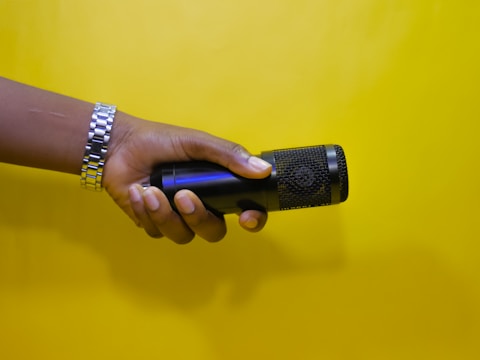 A hand wearing a silver watch is holding a black microphone against a bright yellow background.