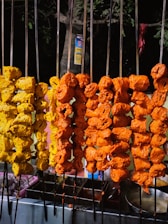 Metal skewers hold marinated chunks of meat, with a rich yellow and red color, suggesting they are being prepared for grilling. The skewers are arranged vertically, likely on a street food stall, with a nighttime setting and a tree trunk visible in the background.