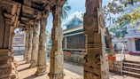 Intricately carved stone pillars with detailed designs are present in a traditional outdoor courtyard. The architecture includes a tiled roof structure with smoke rising, possibly from incense or ritual fires. Palm trees and a clear blue sky are visible in the background, enhancing the serene atmosphere.