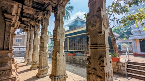 Intricately carved stone pillars with detailed designs are present in a traditional outdoor courtyard. The architecture includes a tiled roof structure with smoke rising, possibly from incense or ritual fires. Palm trees and a clear blue sky are visible in the background, enhancing the serene atmosphere.