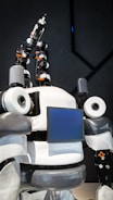 The image features a humanoid robot with mechanical arms and a screen on its chest. The robot has a sleek design with a combination of white, gray, and black colors. It appears to have multiple joints and components, giving it a complex, technical appearance.