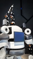 The image features a humanoid robot with mechanical arms and a screen on its chest. The robot has a sleek design with a combination of white, gray, and black colors. It appears to have multiple joints and components, giving it a complex, technical appearance.