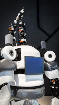 The image features a humanoid robot with mechanical arms and a screen on its chest. The robot has a sleek design with a combination of white, gray, and black colors. It appears to have multiple joints and components, giving it a complex, technical appearance.