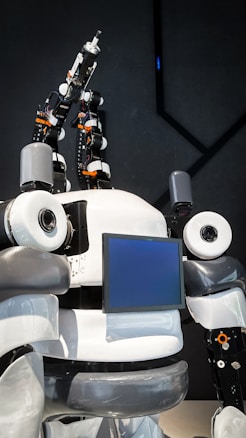 The image features a humanoid robot with mechanical arms and a screen on its chest. The robot has a sleek design with a combination of white, gray, and black colors. It appears to have multiple joints and components, giving it a complex, technical appearance.