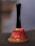 A small, aged handbell with a black handle and a weathered red metallic body. The body shows signs of rust and chipping paint, revealing a worn surface beneath. The text 'Ring for' is partially visible on the bell's body.