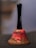 A small, aged handbell with a black handle and a weathered red metallic body. The body shows signs of rust and chipping paint, revealing a worn surface beneath. The text 'Ring for' is partially visible on the bell's body.