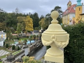 A decorative cream-colored urn sits atop a stone ledge overlooking a formal garden. The garden features manicured hedges, geometric pathways, and classical architecture with colorful facades in the background. Autumn trees with yellow and green foliage are visible, adding to the serene and picturesque setting.