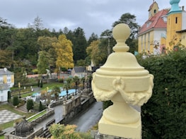 A decorative cream-colored urn sits atop a stone ledge overlooking a formal garden. The garden features manicured hedges, geometric pathways, and classical architecture with colorful facades in the background. Autumn trees with yellow and green foliage are visible, adding to the serene and picturesque setting.