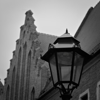 A black and white photograph featuring a historic-looking streetlamp in the foreground. In the background, there is a tall, intricately designed brick building with a steeply pitched roof and gothic-like architectural details.