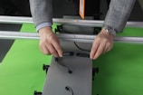 A person wearing a checked coat and a striped shirt is holding and connecting two black cables on a metal frame. The person is wearing a wristwatch, and the background is a bright green surface.
