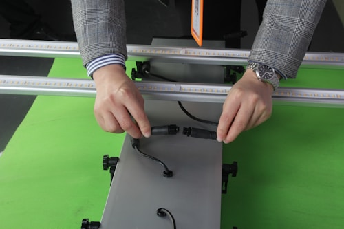 A person wearing a checked coat and a striped shirt is holding and connecting two black cables on a metal frame. The person is wearing a wristwatch, and the background is a bright green surface.