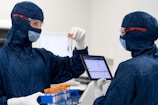 Two individuals are wearing blue protective suits, masks, and goggles in a sterile laboratory environment. One is holding a test tube with an orange cap, while the other holds a digital tablet displaying information. A tray with other test tubes is visible, indicating they are involved in some form of scientific or medical research.