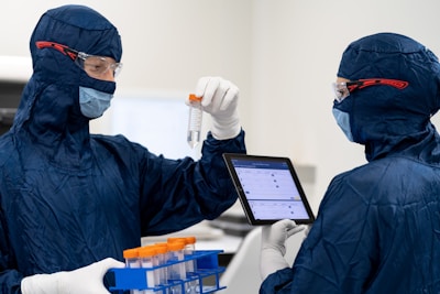 Two individuals are wearing blue protective suits, masks, and goggles in a sterile laboratory environment. One is holding a test tube with an orange cap, while the other holds a digital tablet displaying information. A tray with other test tubes is visible, indicating they are involved in some form of scientific or medical research.