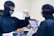 Two individuals are wearing blue protective suits, masks, and goggles in a sterile laboratory environment. One is holding a test tube with an orange cap, while the other holds a digital tablet displaying information. A tray with other test tubes is visible, indicating they are involved in some form of scientific or medical research.