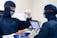 Two individuals are wearing blue protective suits, masks, and goggles in a sterile laboratory environment. One is holding a test tube with an orange cap, while the other holds a digital tablet displaying information. A tray with other test tubes is visible, indicating they are involved in some form of scientific or medical research.