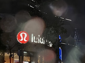 A dimly lit night scene with a store featuring a bright red circular logo with a white design. The store's name is partially visible next to the logo. The foreground includes some blurred foliage, and there are lights from a nearby building in the background, adding to the urban atmosphere.