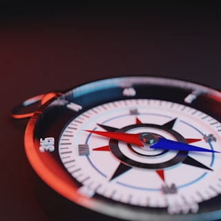 A close-up view of a compass lying on a dark surface, highlighting the compass rose with clear red and blue needles pointing in opposite directions. The numbers and cardinal directions are visible along the edges.