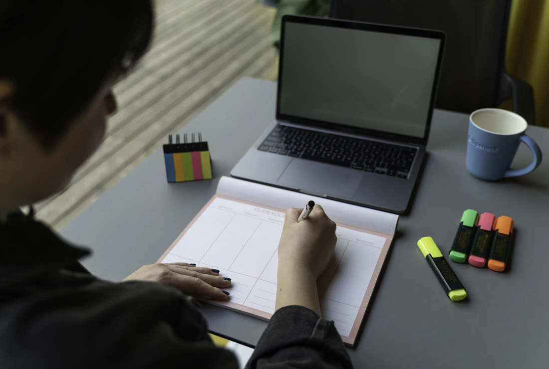 Pessoa de costas pesquisando no laptop e anotando em um caderno