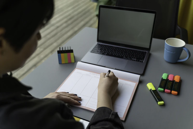 Pessoa de costas pesquisando no laptop e anotando em um caderno