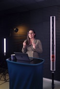 A woman is standing at a small podium with a laptop in front of her. She is wearing glasses and is smiling. The setting includes soft lighting with acoustic panels on the walls, suggesting a professional or recording environment.