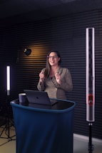 A woman is standing at a small podium with a laptop in front of her. She is wearing glasses and is smiling. The setting includes soft lighting with acoustic panels on the walls, suggesting a professional or recording environment.