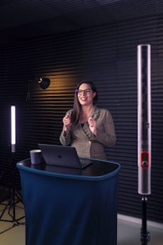 A woman is standing at a small podium with a laptop in front of her. She is wearing glasses and is smiling. The setting includes soft lighting with acoustic panels on the walls, suggesting a professional or recording environment.