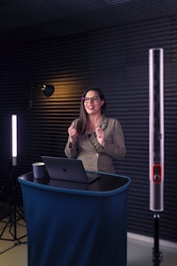 A woman is standing at a small podium with a laptop in front of her. She is wearing glasses and is smiling. The setting includes soft lighting with acoustic panels on the walls, suggesting a professional or recording environment.