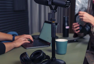 A podcast setup with a large microphone on a stand positioned on a table. Nearby are a pair of black headphones and a laptop. Two people are in the background, one typing on the laptop and the other holding headphones. The setting appears to be a studio with soundproofing elements.