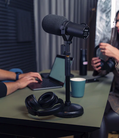 A podcast setup with a large microphone on a stand positioned on a table. Nearby are a pair of black headphones and a laptop. Two people are in the background, one typing on the laptop and the other holding headphones. The setting appears to be a studio with soundproofing elements.