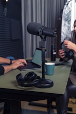 A podcast setup with a large microphone on a stand positioned on a table. Nearby are a pair of black headphones and a laptop. Two people are in the background, one typing on the laptop and the other holding headphones. The setting appears to be a studio with soundproofing elements.