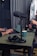A podcast setup with a large microphone on a stand positioned on a table. Nearby are a pair of black headphones and a laptop. Two people are in the background, one typing on the laptop and the other holding headphones. The setting appears to be a studio with soundproofing elements.