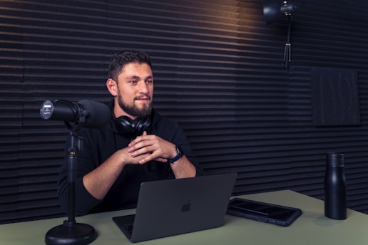 A person sits at a table in a soundproofed room, speaking into a large microphone. They have headphones around their neck and a laptop open in front of them. The room is equipped with professional audio recording equipment, and there is a water bottle on the table.