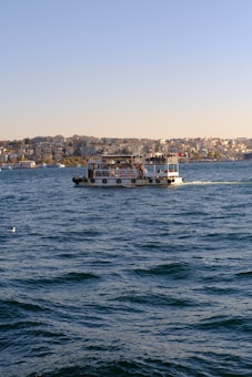 A passenger boat is sailing on a large body of water with gentle waves. In the distance, a densely built urban landscape fills the background with multiple buildings under a clear blue sky.