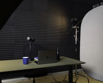 A podcast or recording setup featuring a table with a laptop, a microphone on a stand, a blue coffee mug, and a black bottle. The backdrop is lined with acoustic foam for sound insulation, and there is a large light diffuser on a stand.