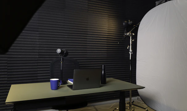 A podcast or recording setup featuring a table with a laptop, a microphone on a stand, a blue coffee mug, and a black bottle. The backdrop is lined with acoustic foam for sound insulation, and there is a large light diffuser on a stand.
