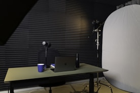 A podcast or recording setup featuring a table with a laptop, a microphone on a stand, a blue coffee mug, and a black bottle. The backdrop is lined with acoustic foam for sound insulation, and there is a large light diffuser on a stand.