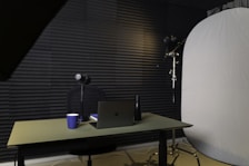 A podcast or recording setup featuring a table with a laptop, a microphone on a stand, a blue coffee mug, and a black bottle. The backdrop is lined with acoustic foam for sound insulation, and there is a large light diffuser on a stand.