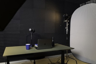 A podcast or recording setup featuring a table with a laptop, a microphone on a stand, a blue coffee mug, and a black bottle. The backdrop is lined with acoustic foam for sound insulation, and there is a large light diffuser on a stand.