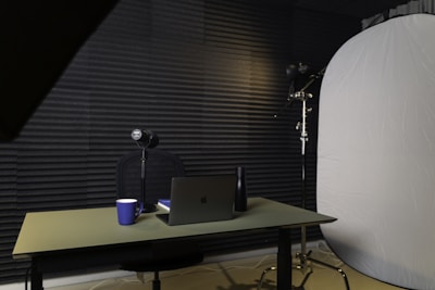 A podcast or recording setup featuring a table with a laptop, a microphone on a stand, a blue coffee mug, and a black bottle. The backdrop is lined with acoustic foam for sound insulation, and there is a large light diffuser on a stand.