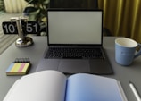 A well-organized workspace featuring an open laptop with a blank screen, a blue mug, a flip clock displaying the time 10:51, a notebook with colored sticky note tabs, and an open notebook with a pen.