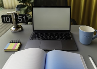 A well-organized workspace featuring an open laptop with a blank screen, a blue mug, a flip clock displaying the time 10:51, a notebook with colored sticky note tabs, and an open notebook with a pen.