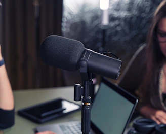 Close-up of a podcast or recording setup featuring a large microphone at the center with two people in the background. One is a woman with glasses, and the other is a person on the left partially out of frame. A laptop and a tablet are on the table.