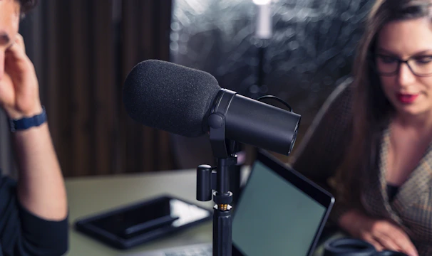 Close-up of a podcast or recording setup featuring a large microphone at the center with two people in the background. One is a woman with glasses, and the other is a person on the left partially out of frame. A laptop and a tablet are on the table.