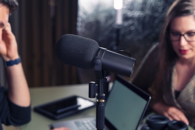 Close-up of a podcast or recording setup featuring a large microphone at the center with two people in the background. One is a woman with glasses, and the other is a person on the left partially out of frame. A laptop and a tablet are on the table.